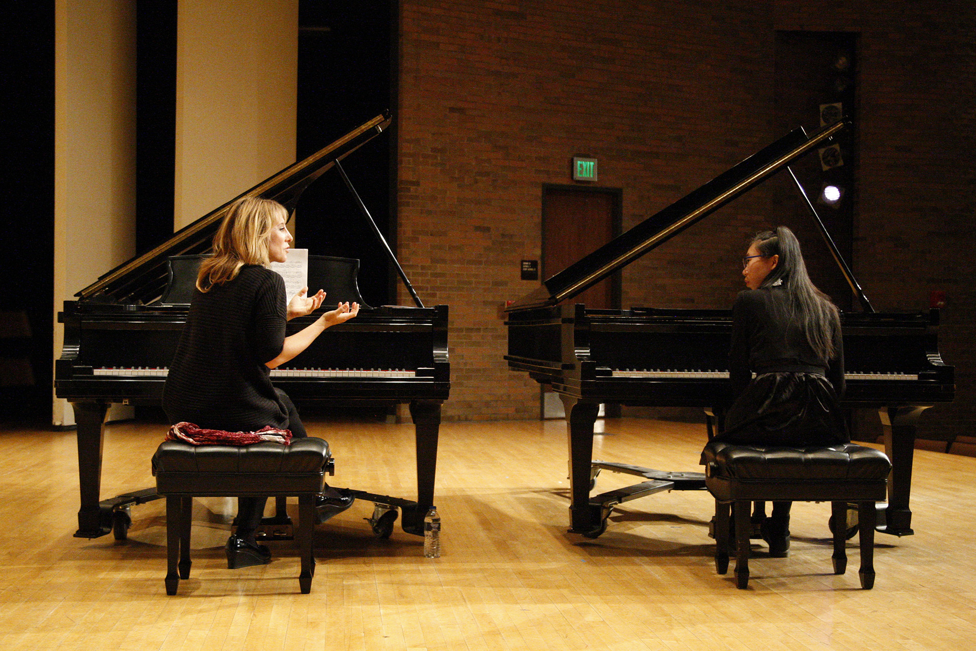 Two women talking while sitting at two separate pianos on stage