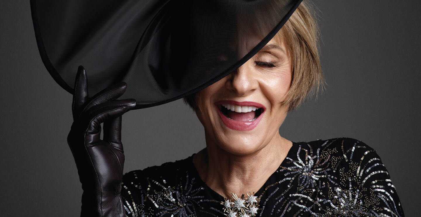 patti lupone laughs while holding a wide-brimmed hat